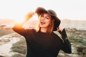 beautiful blonde young woman in glasses and holding hat and winking on the beach with sunset