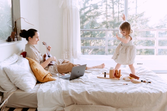 A mom resting in bed while her daughter is dancing on it.